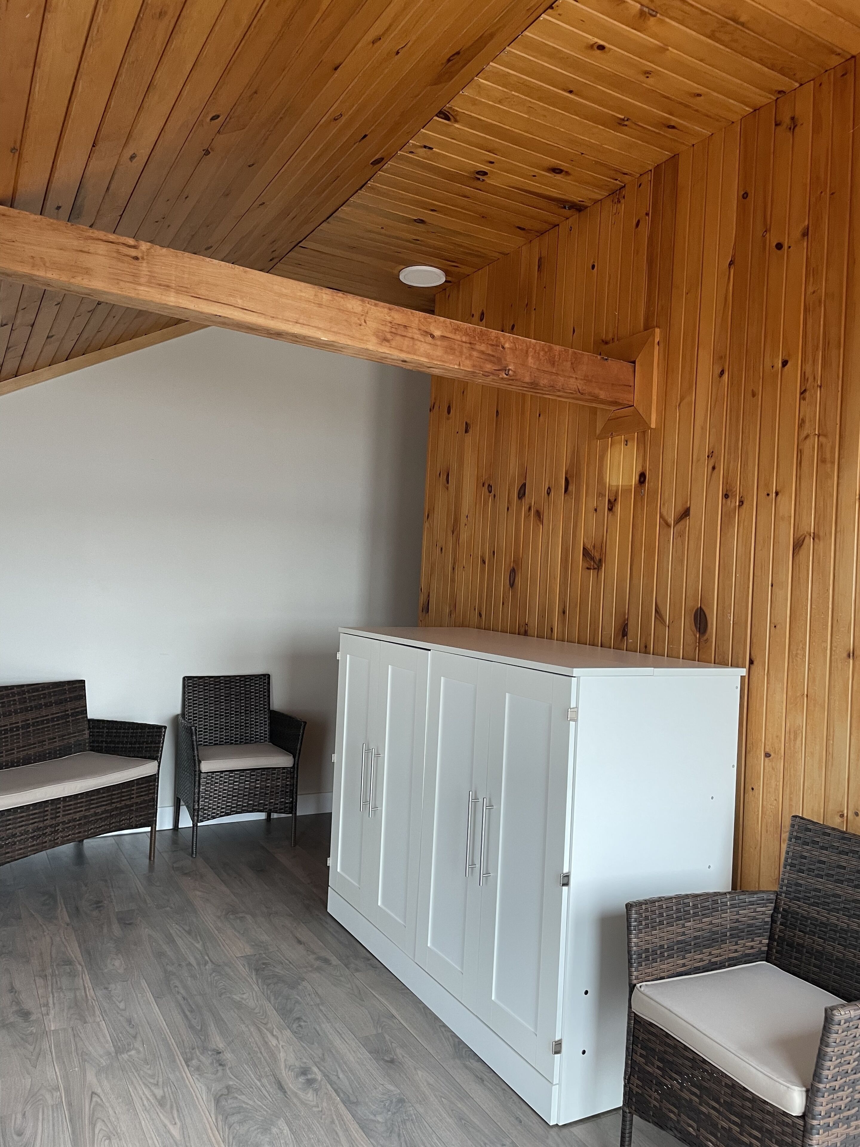 A room with a wooden ceiling, a white wardrobe, and wicker furniture.