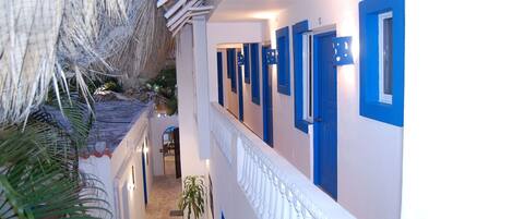 Un pasillo de hotel con puertas azules, barandilla blanca y techo de paja.