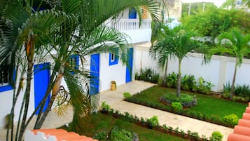 Un patio con un edificio blanco, con puertas y ventanas azules, rodeado de palmeras y un jardín bien cuidado.