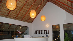 A reception area with a curved counter, hanging wicker lamps, a thatched ceiling, and large windows. - Hotel Playa Catalina (La Romana)