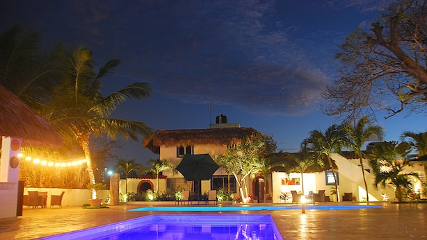 A pool area with a thatched-roof structure, palm trees, and a building with a balcony. - Hotel Playa Catalina (La Romana)