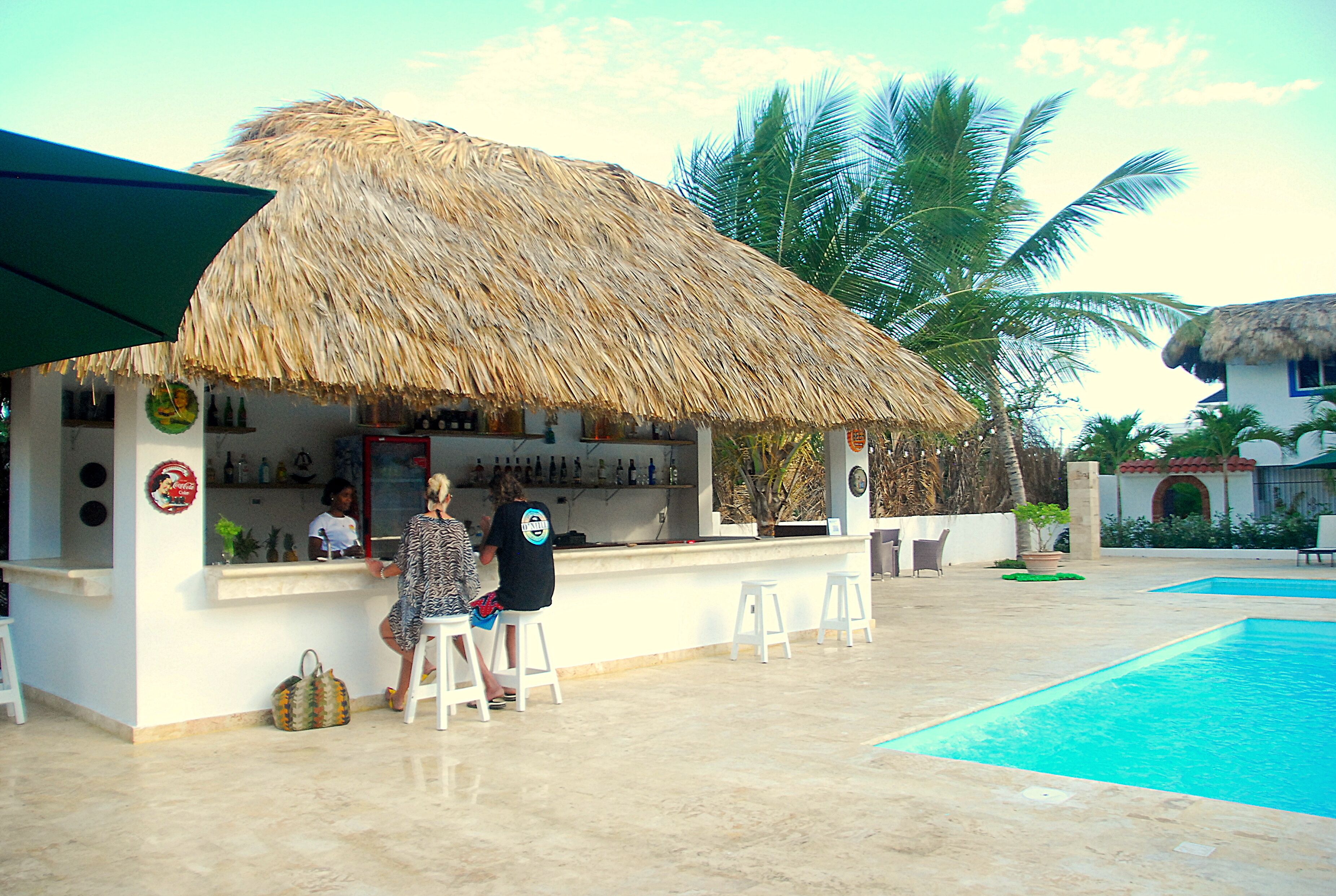 A bar area with a thatched roof, white walls, and a pool in the background.