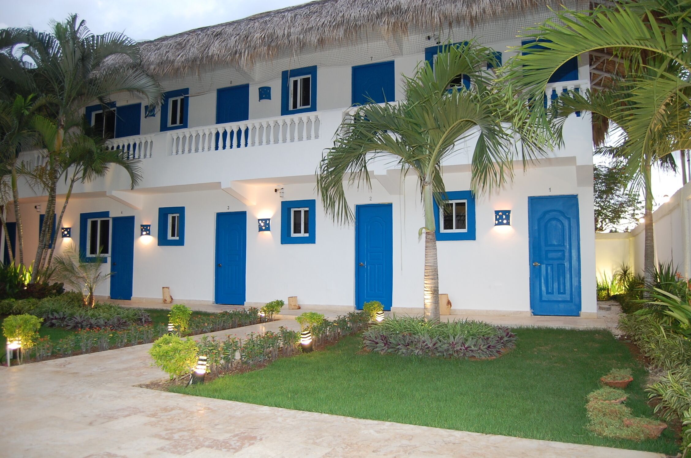 Un edificio de varios pisos con puertas y ventanas azules, rodeado de jardines cuidados y palmeras.