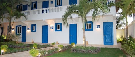 Un edificio de varios pisos con puertas y ventanas azules, rodeado de jardines cuidados y palmeras.