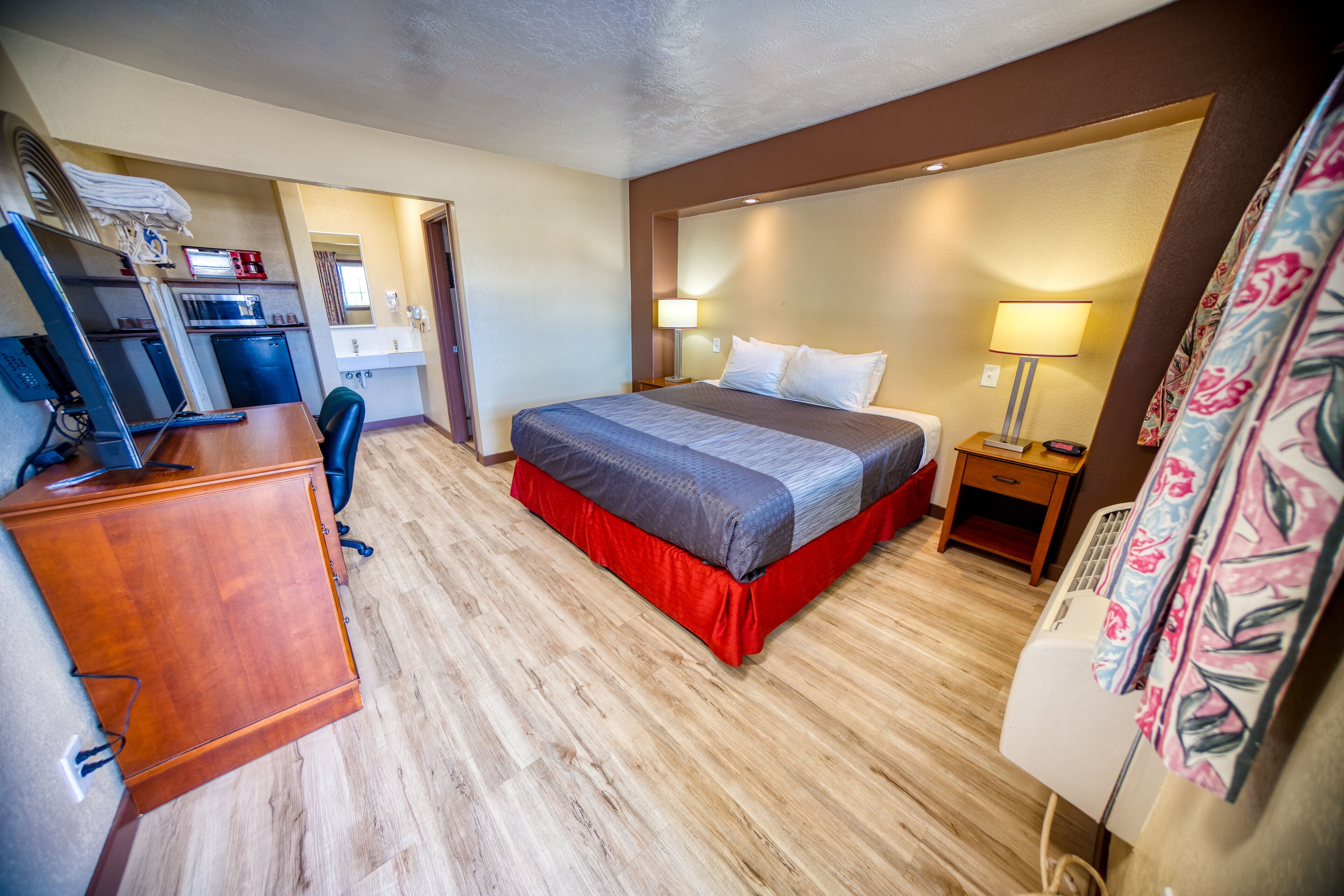 A hotel room with a bed, desk, chair, and a small table with a lamp.