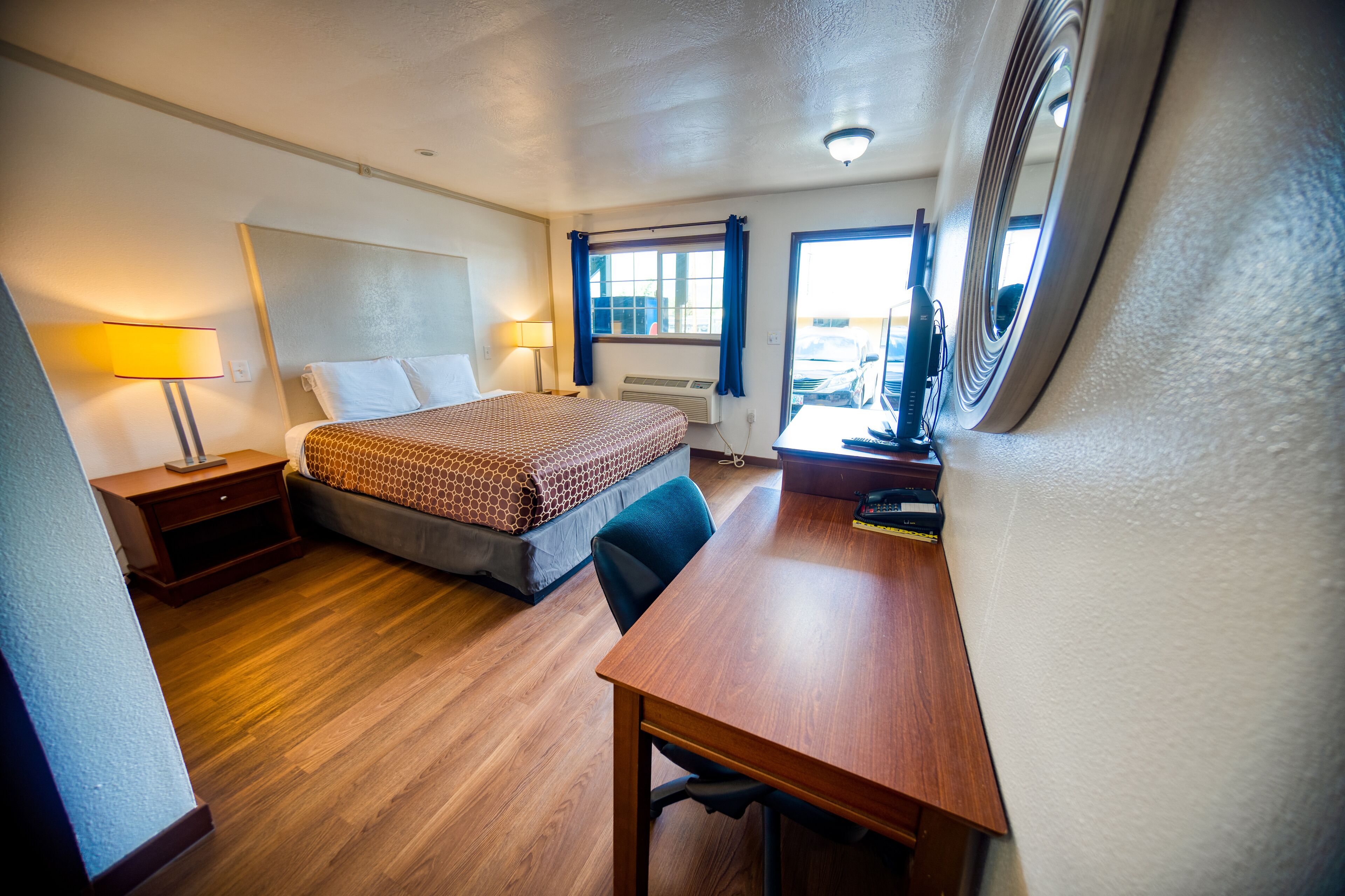 A hotel room with a bed, bedside table, desk, chair, lamp, and a window with curtains.