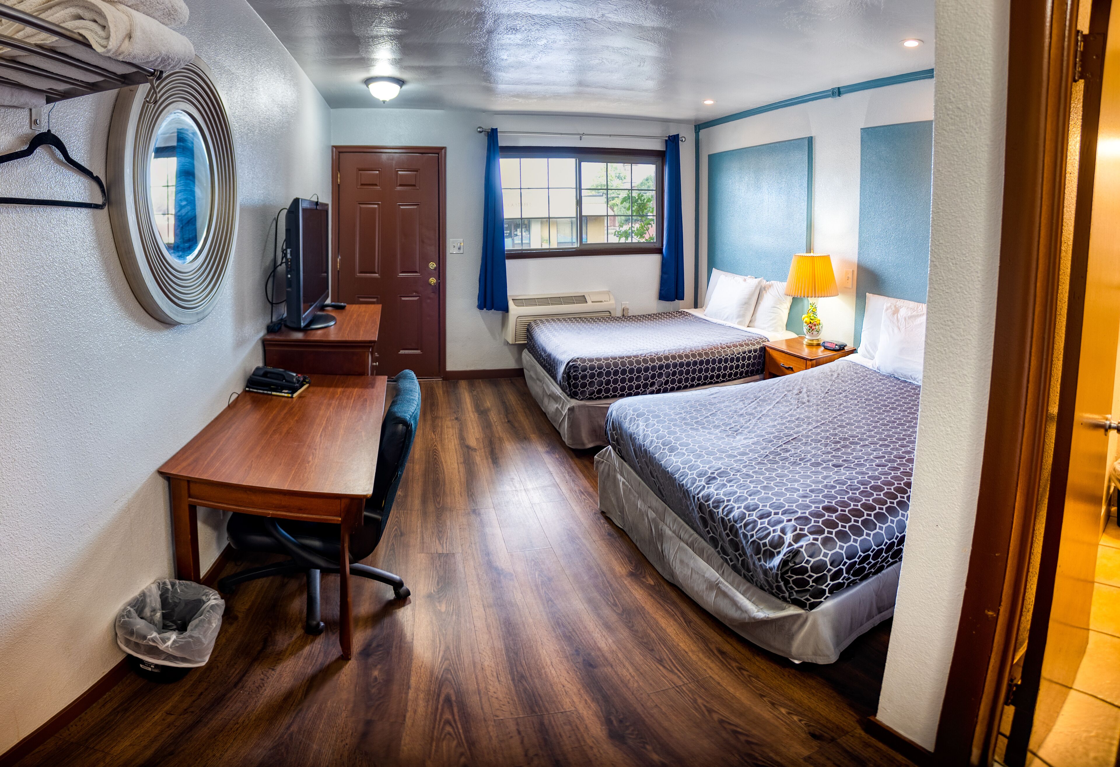 A compact hotel room with a wooden desk, two beds, a TV, and a window with curtains.