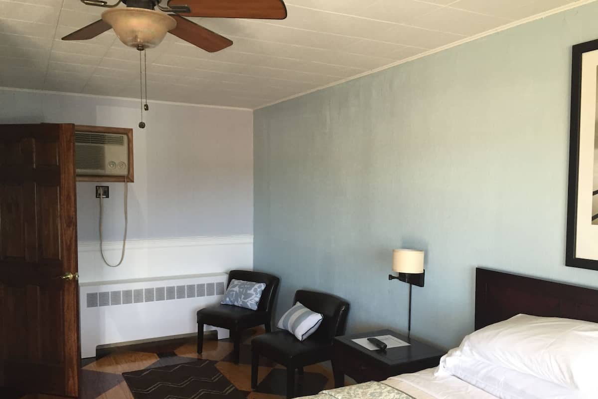 A hotel room with a bed, two chairs, a nightstand, a lamp, a ceiling fan, and a door leading to a bathroom.