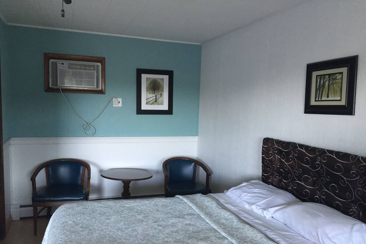 A bedroom with a bed, two chairs, a small table, and two framed pictures on the wall.