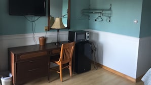 Quarto com paredes verde-água, uma escrivaninha de madeira, uma cadeira, uma televisão, um abajur, um micro-ondas e um porta-roupas.
