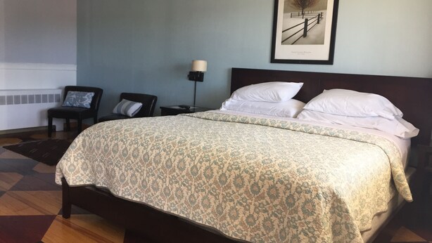 Cama bem arrumada com colcha estampada, quadro pendurado acima, cadeira, abajur e chão xadrez.