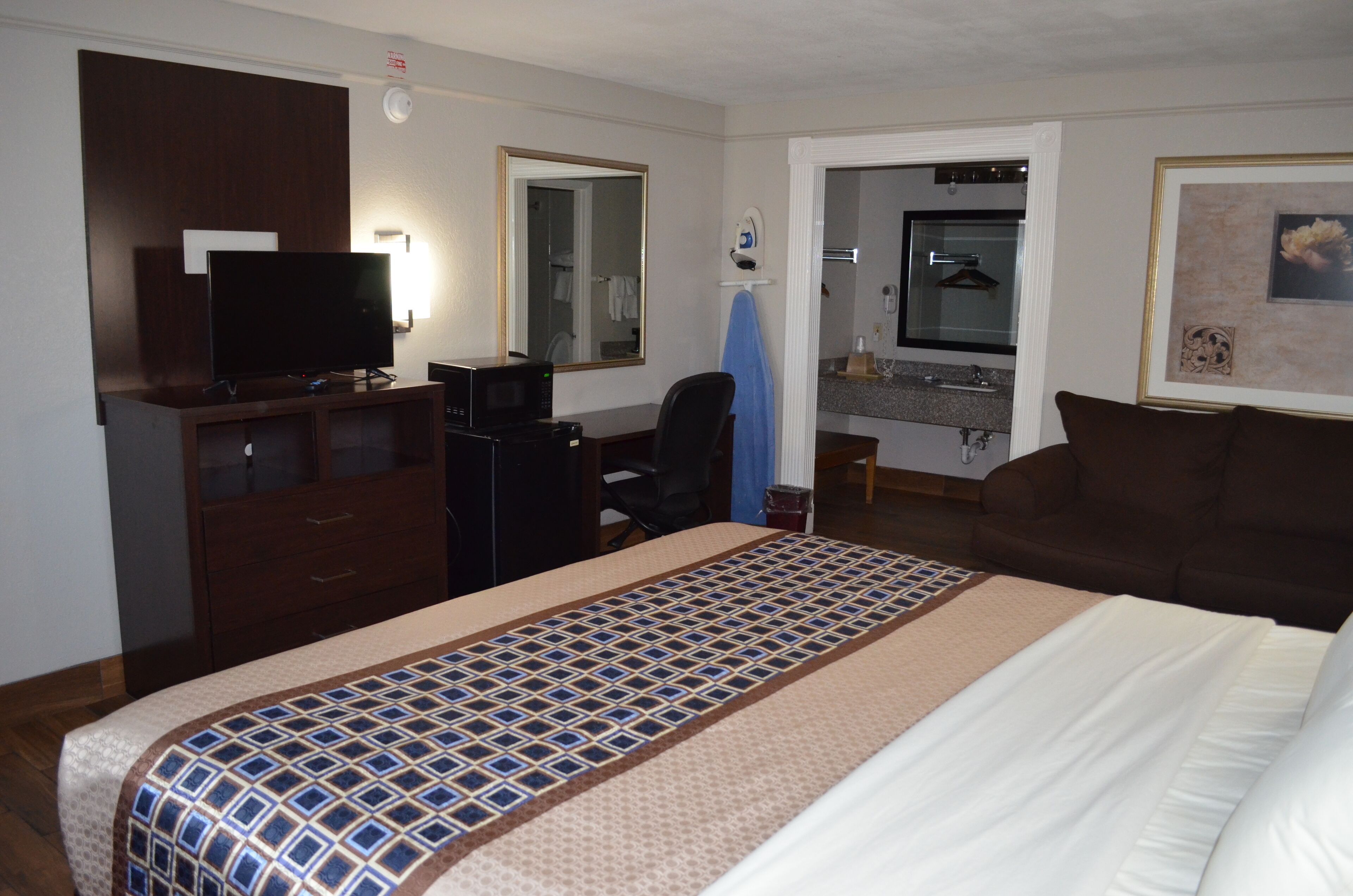 Une chambre d’hôtel comprenant un lit, un bureau avec un ordinateur, une télévision, un miroir, une salle de bain avec un lavabo et un porte-serviettes, et un tableau encadré accroché au mur.