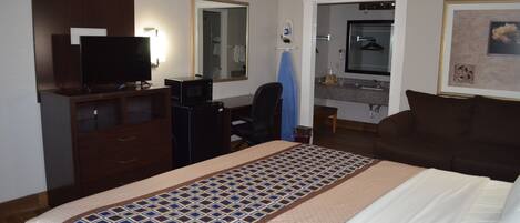 A hotel room with a bed, a desk with a computer, a television, a mirror, a bathroom with a sink and towel rack, and a framed picture on the wall.