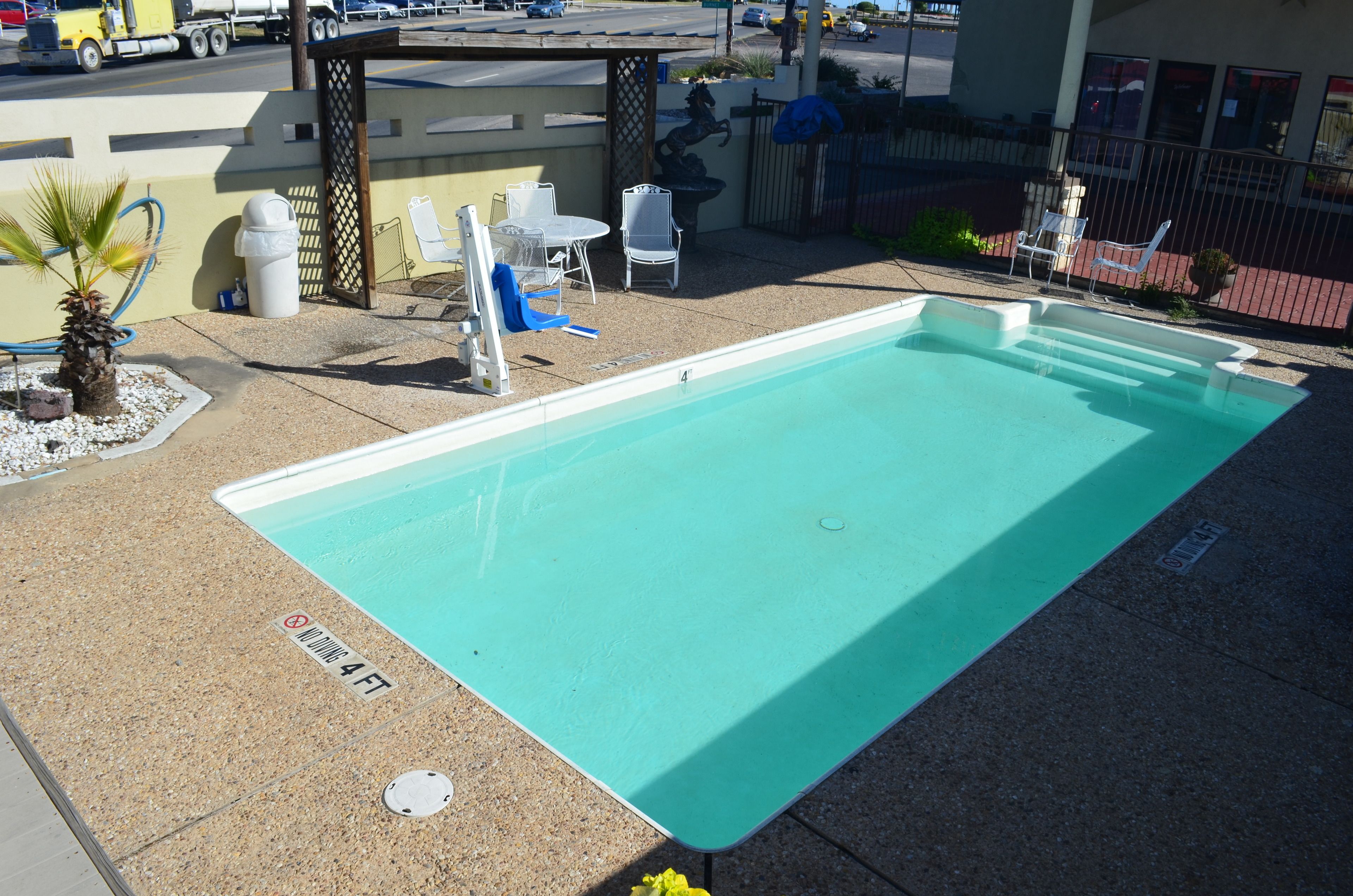 Una piscina rectangular con un cartel de prohibición de saltos, rodeada por una terraza con sillas y una pequeña palmera.