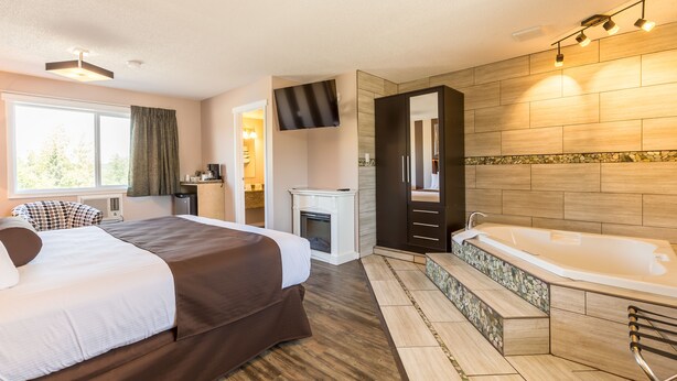 A modern hotel room with a large bed, a separate bathroom with a bathtub, a fireplace, and a view of the outdoors. - Carmel Inn (Prince George)