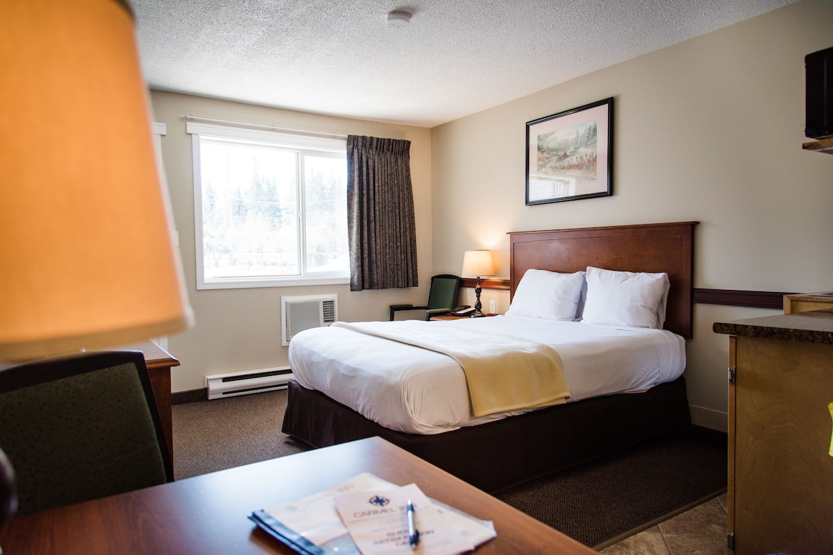 Une chambre d’hôtel avec un lit, un bureau, une chaise, une lampe, un tableau au mur et une fenêtre avec des rideaux.