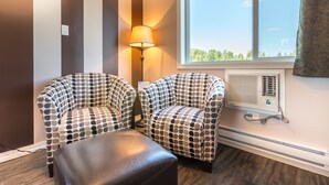 A room with two patterned armchairs, a small ottoman, a standing lamp, and a window with a view of greenery. - Carmel Inn (Prince George)