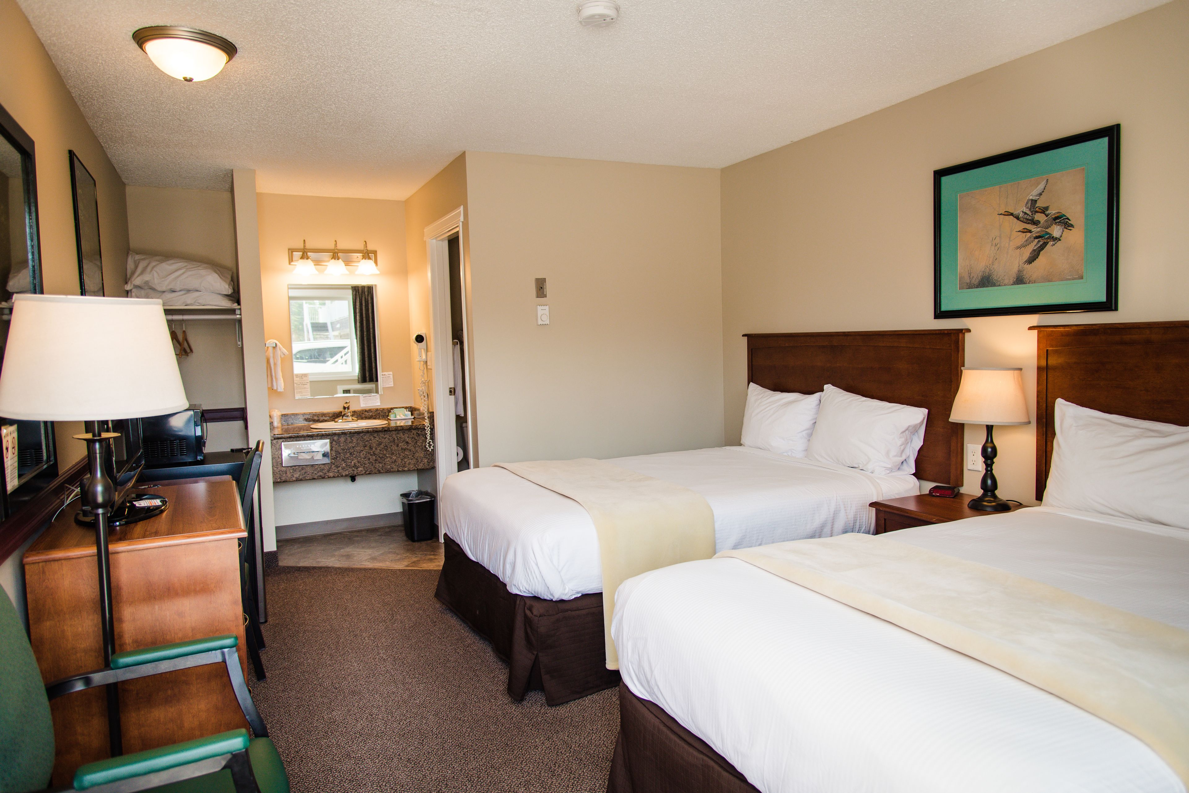 Une chambre d’hôtel avec deux lits, un bureau, une chaise, une lampe, un miroir, un lavabo et un tableau encadré au mur.