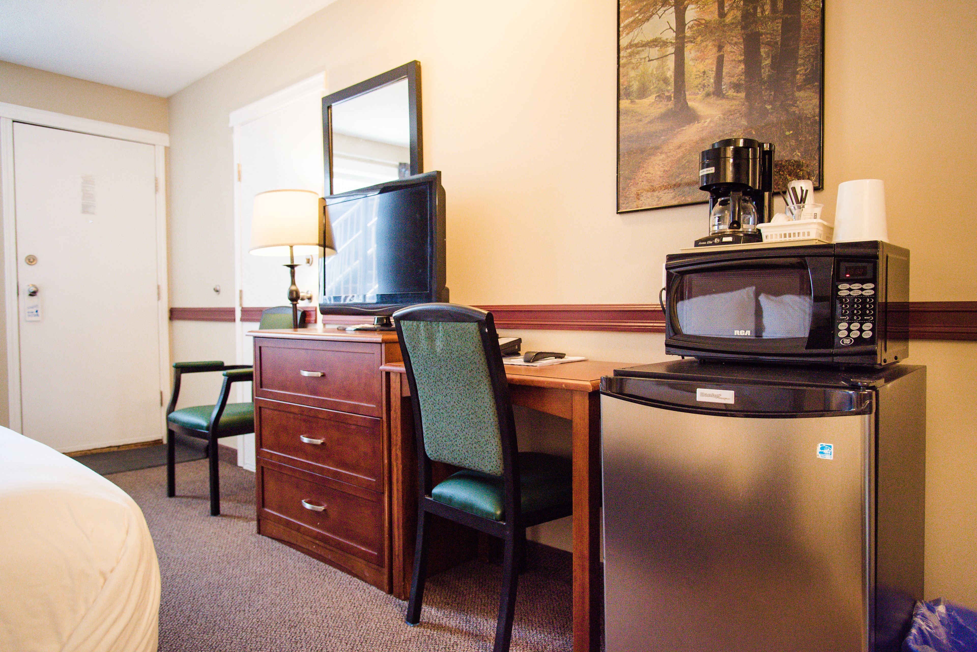 Une chambre d’hôtel comprenant un lit, un bureau avec un ordinateur, une chaise, un réfrigérateur, un four à micro-ondes, une machine à café et un tableau accroché au mur.