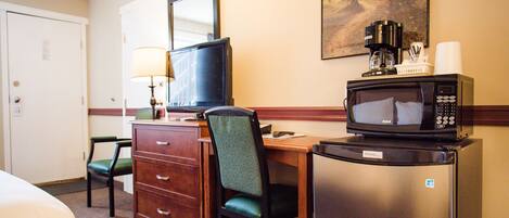 Une chambre d’hôtel comprenant un lit, un bureau avec un ordinateur, une chaise, un réfrigérateur, un four à micro-ondes, une machine à café et un tableau accroché au mur.
