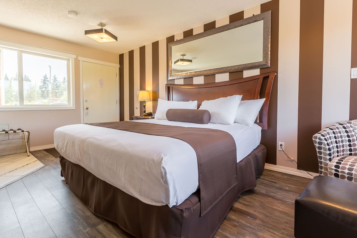 Une chambre d’hôtel avec un grand lit, une tête de lit en bois, une fenêtre donnant sur la verdure et un fauteuil à motifs.
