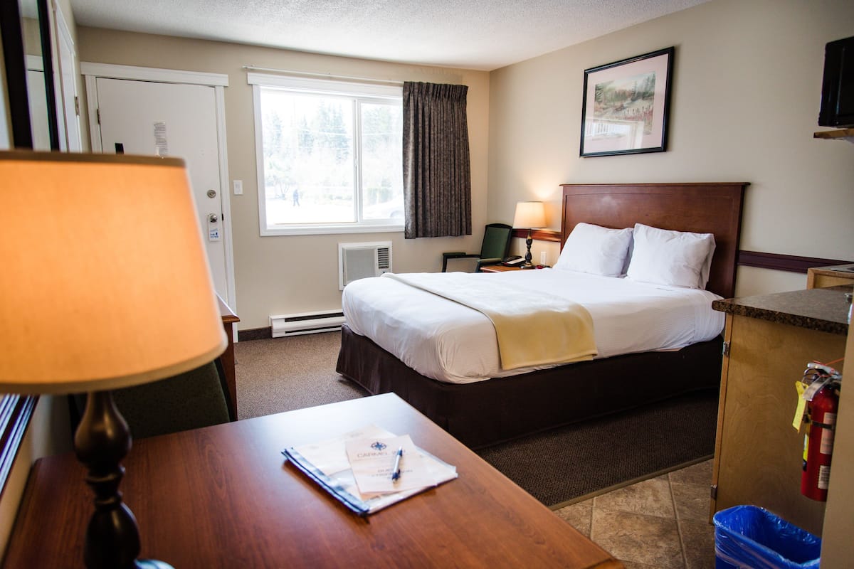 Une chambre d’hôtel avec un lit, un bureau avec une lampe, une chaise, une fenêtre avec des rideaux, un tableau au mur et un extincteur.