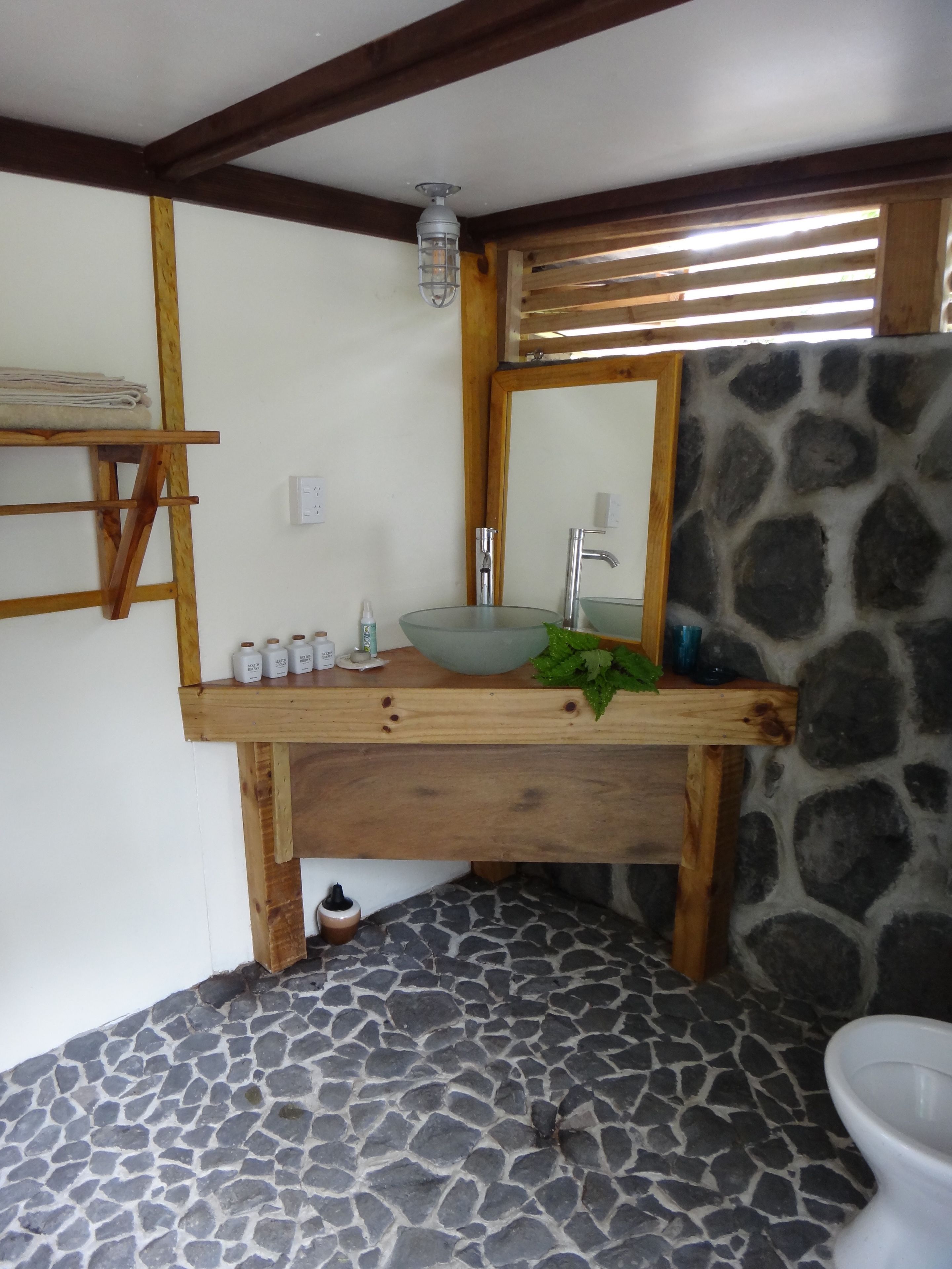 A bathroom with a stone floor, a wooden vanity with a glass bowl sink, a mirror, and a toilet.