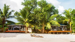 Due cabine sulla spiaggia con verande aperte, circondate da palme e una spiaggia sabbiosa.