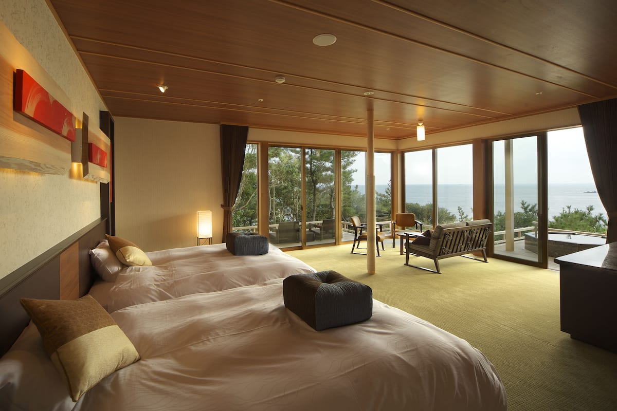 A modern hotel room with two large beds, a balcony with a view of the sea, and a dining area with a table and chairs.
