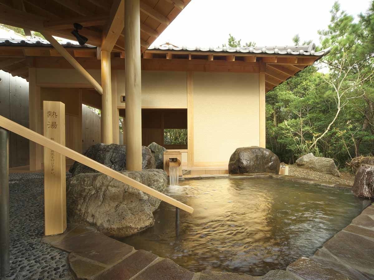 Un onsen japonés tradicional con un paseo de madera, rocas grandes y una estructura de techo con tejas.