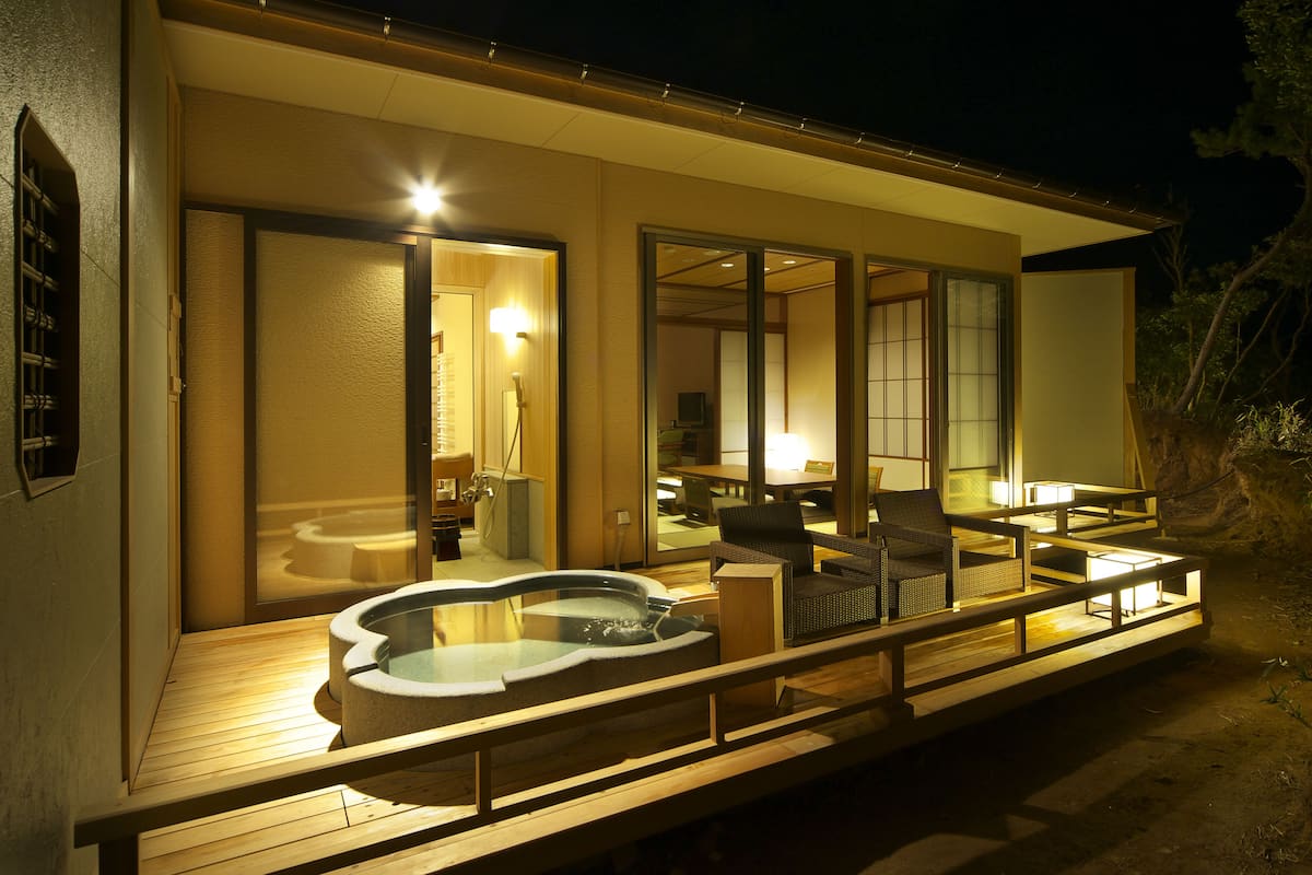 A modern building with a hot tub on the deck, wooden furniture, and a well-lit interior.