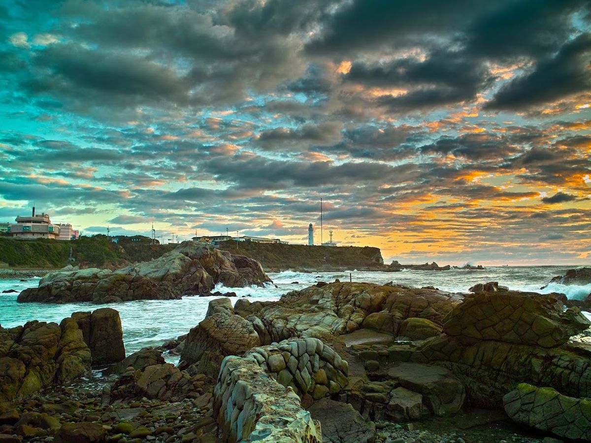 A rocky coastal landscape with a lighthouse and buildings in the background.
