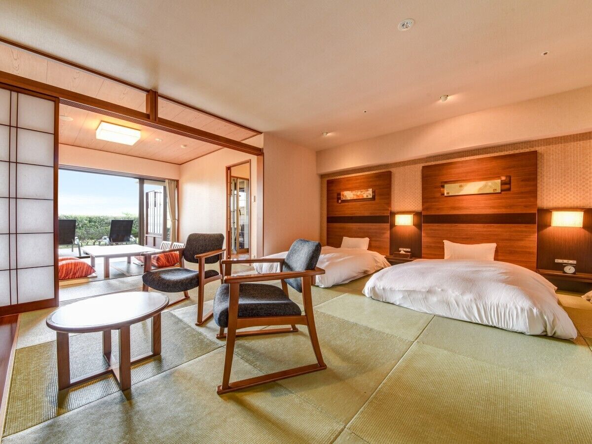 Ein Hotelzimmer mit Tatami-Boden, einem Bett, einer Sitzecke mit Stühlen, einem Tisch und Blick durch ein großes Fenster.