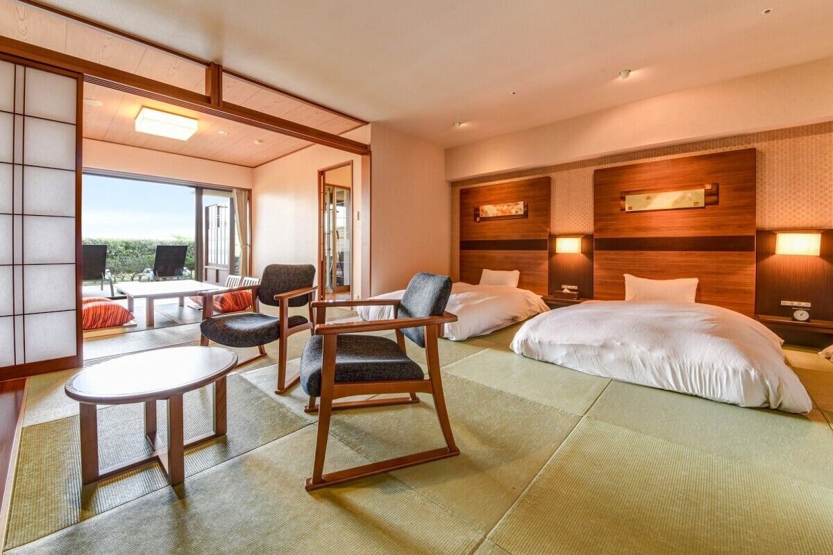 Habitación de hotel con suelo de tatami, una cama, zona de estar con sillas, una mesa y vistas a través de un ventanal grande.