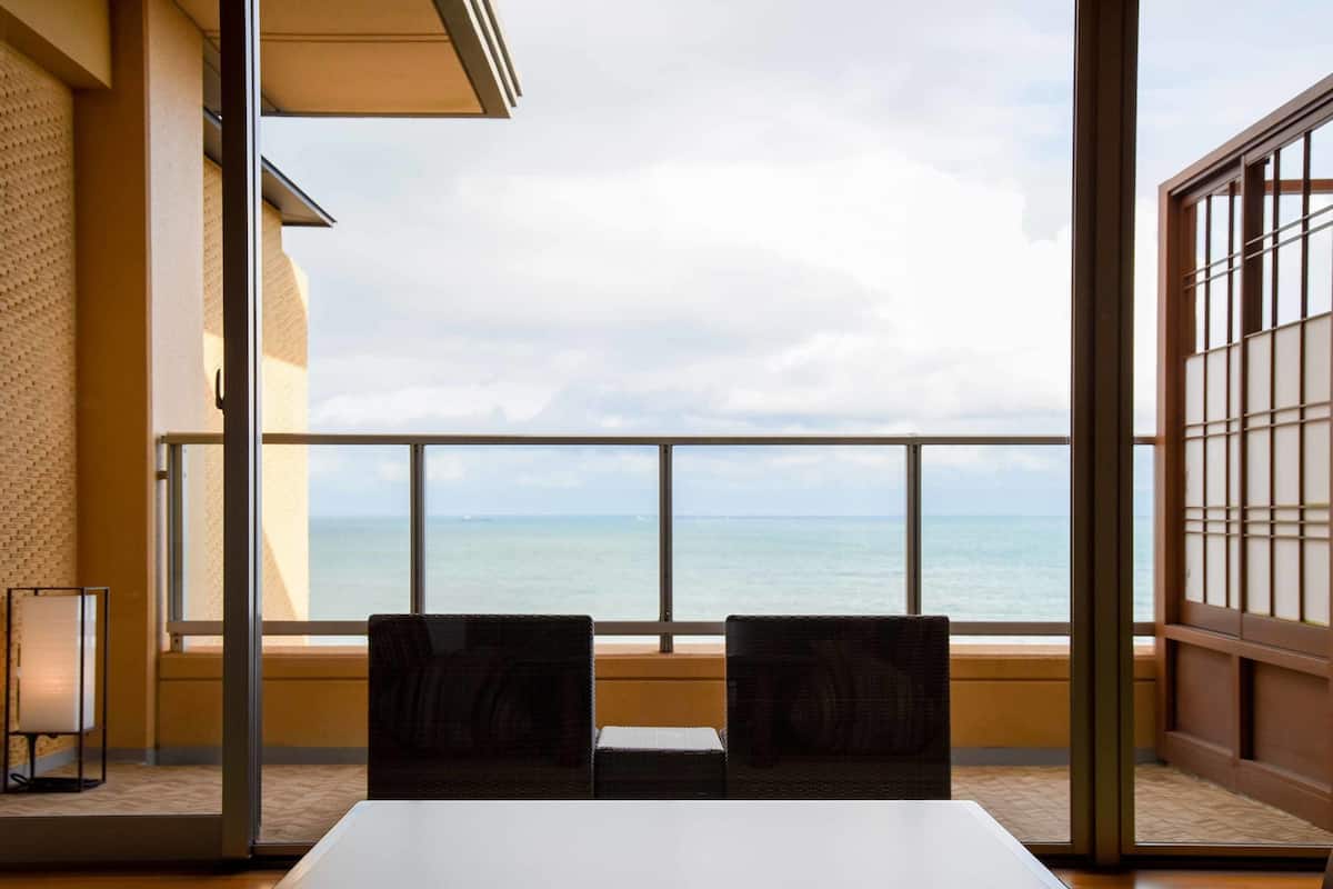 A room with a large window overlooking the sea, two chairs, and a table.