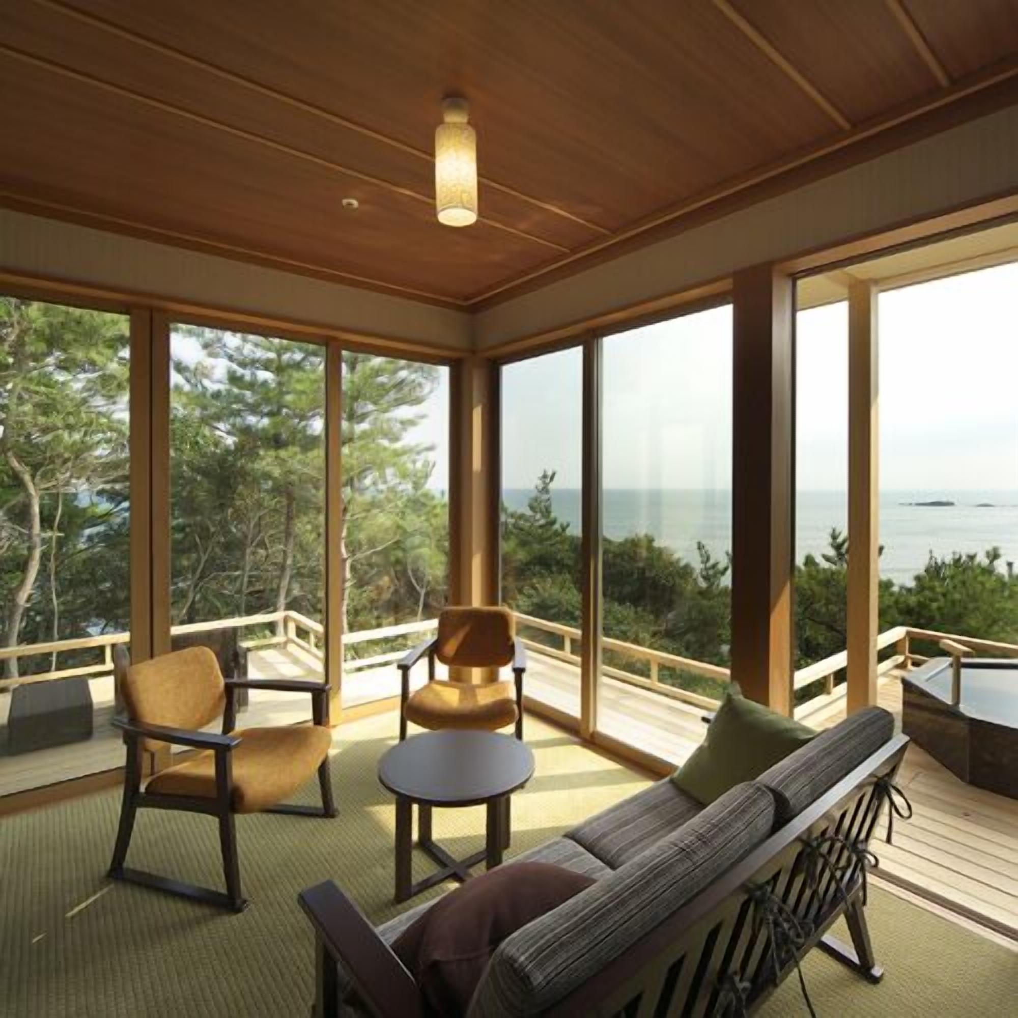 Une pièce spacieuse dotée d’une grande fenêtre donnant sur la mer, d’un plafond en bois et d’un coin salon avec un canapé et des fauteuils.