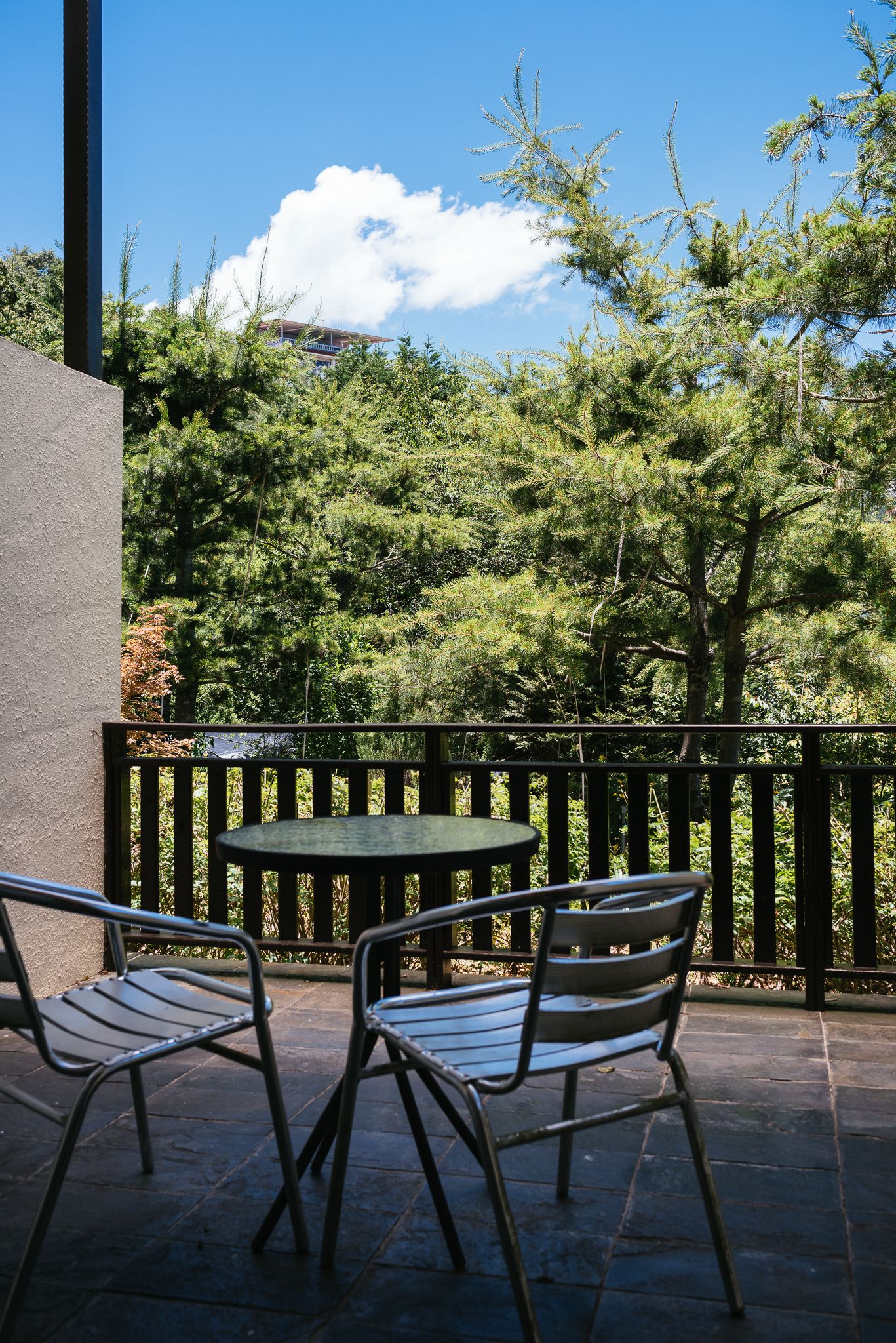 An outdoor patio with a round table and two chairs, surrounded by a wooden railing, overlooking a forested area.