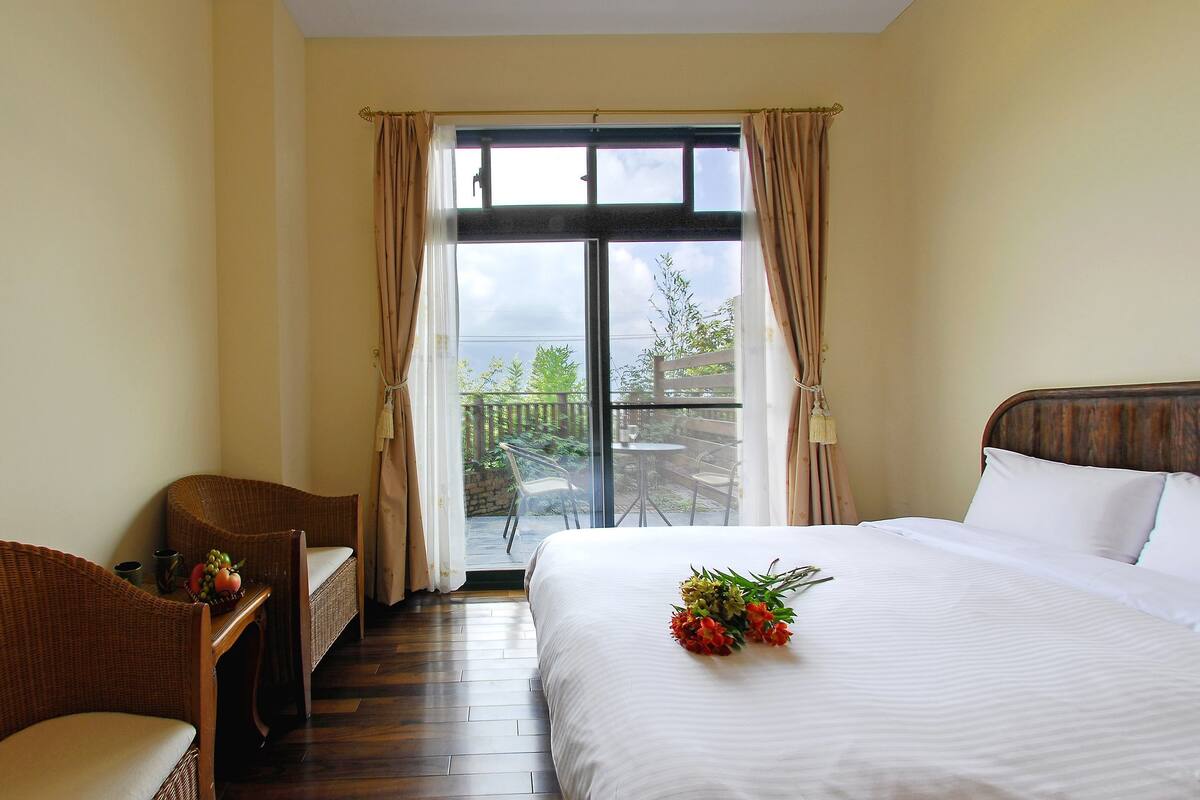 A hotel room with a bed, two wicker chairs, a small table with fruit, and a balcony with a table and chairs.