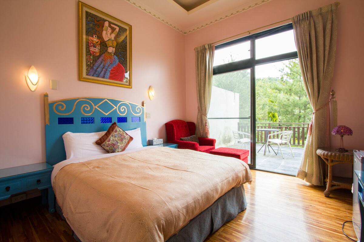 A bedroom with a large bed, a red chair, a balcony with a table and chairs, and a painting on the wall.