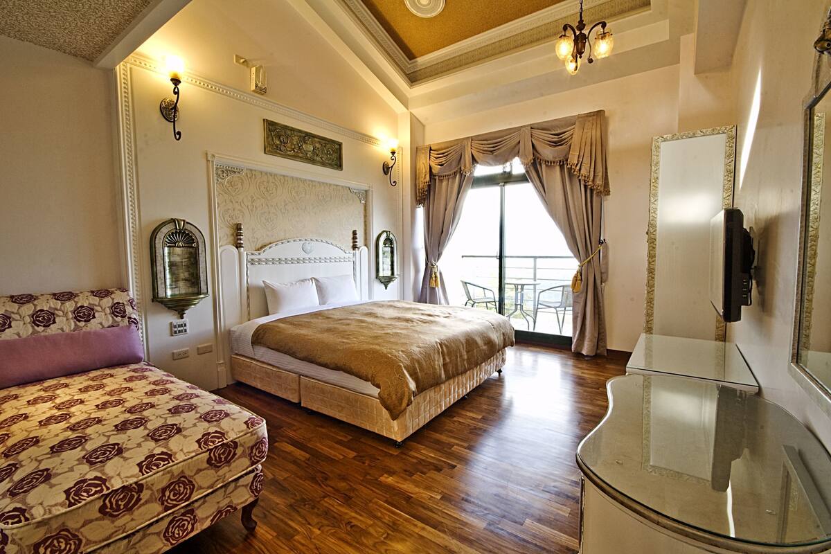 A bedroom with a large bed, a bedside table, a chair, and a balcony with a view.