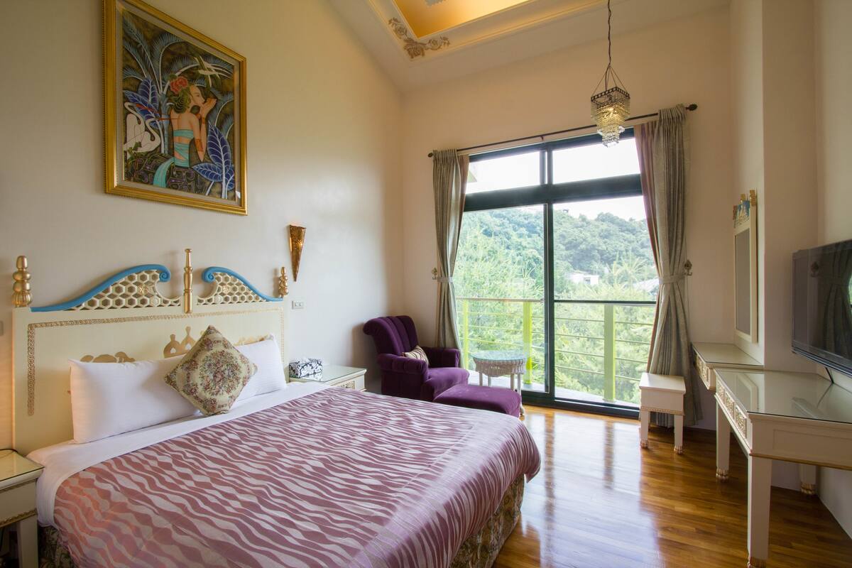 A bedroom with a bed, a chair, a desk, and a large window with a view of greenery.