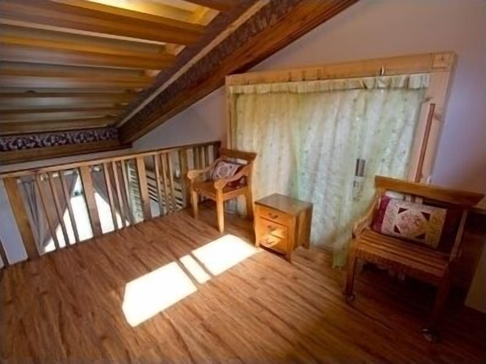 Una habitación con piso de madera, techo inclinado, una ventana con cortinas y una pequeña mesa de madera con una silla.