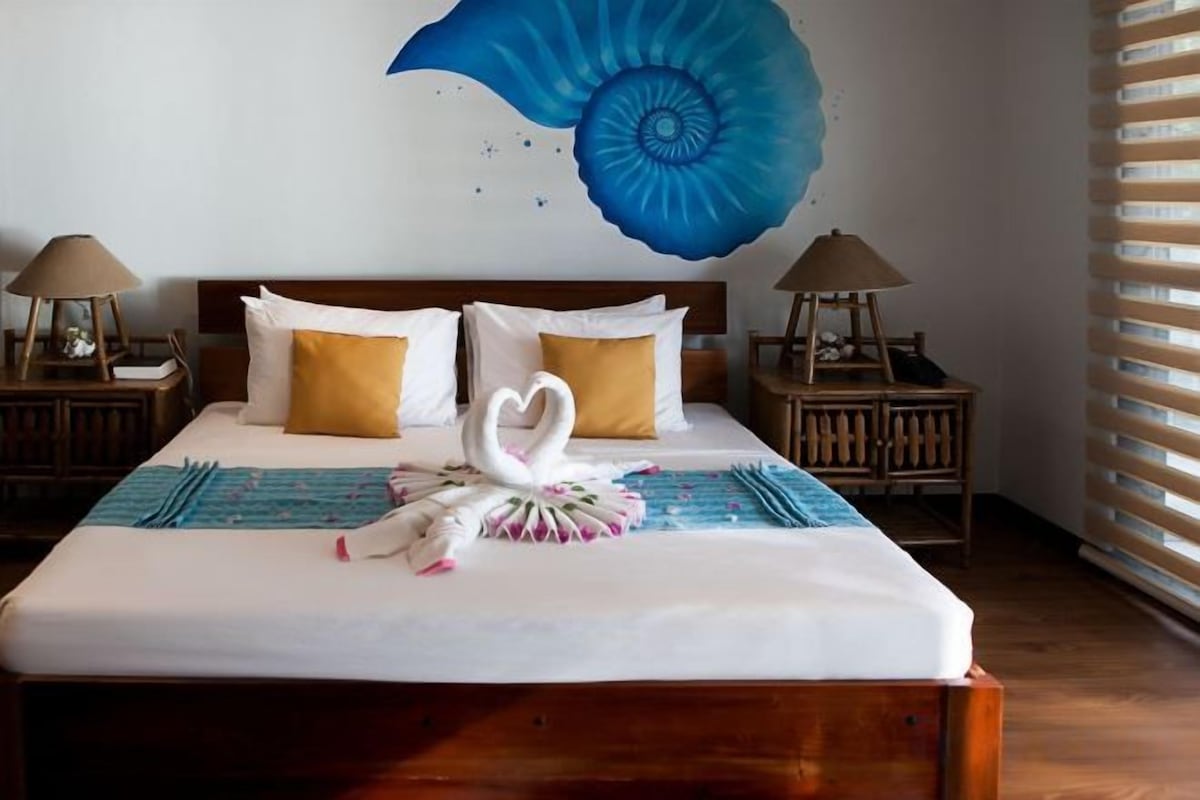 Un lit bien fait, agrémenté d’un coussin décoratif en forme de cygne, une œuvre d’art représentant une coquille bleue accrochée au mur et une tête de lit en bois.