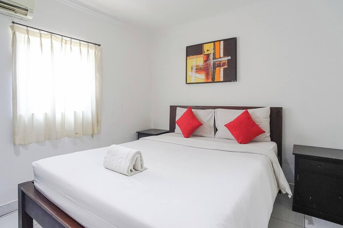 A neatly made bed with white bedding and a rolled towel, a nightstand with a lamp, a painting on the wall, and a window with curtains.