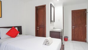 Ett hotellrum med en säng, en röd kudde, en vit handduk, ett handfat och två trädörrar.