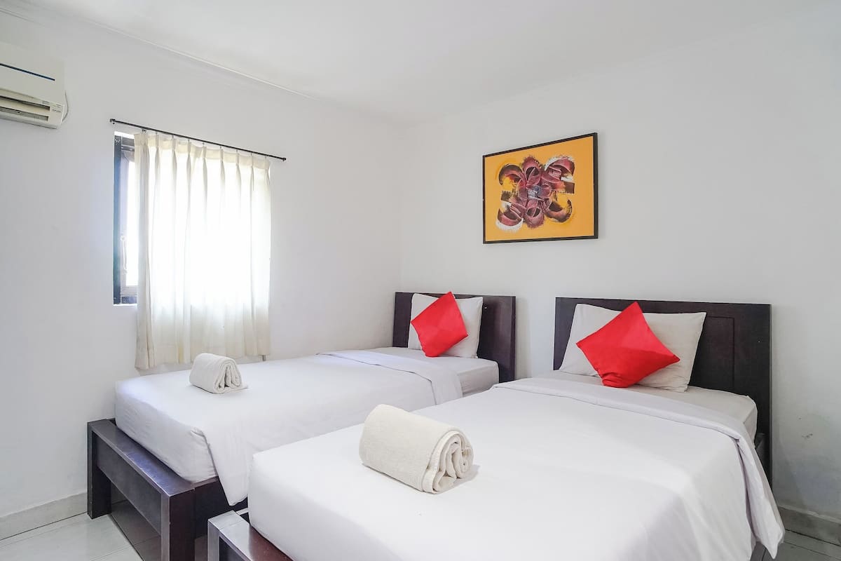 A hotel room with two beds, each with white linens and red pillows, a large window with sheer curtains, and a framed artwork on the wall.