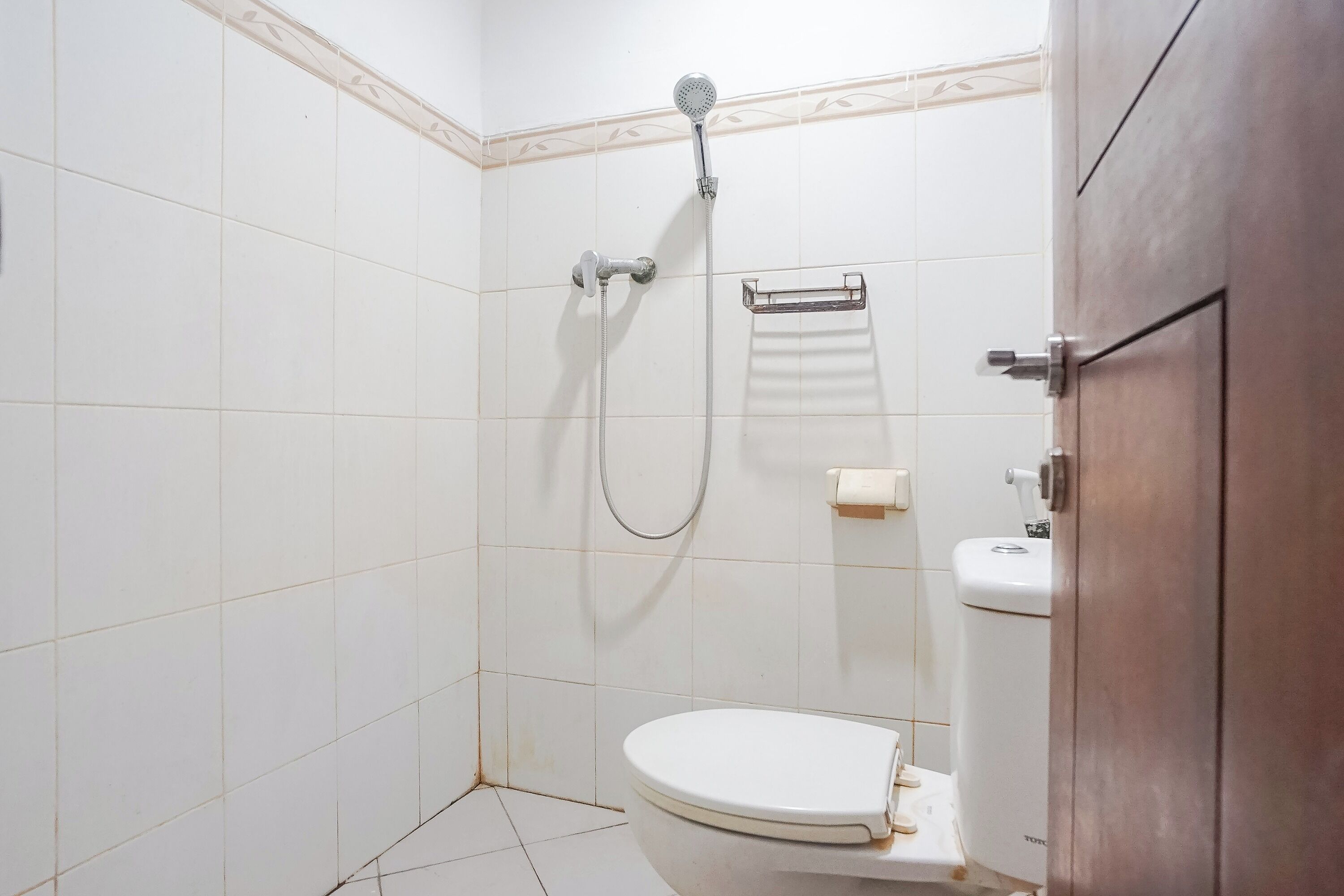 Un baño pequeño y limpio con azulejos blancos, un inodoro, una ducha con cabezal de ducha manual y un perchero para toallas.