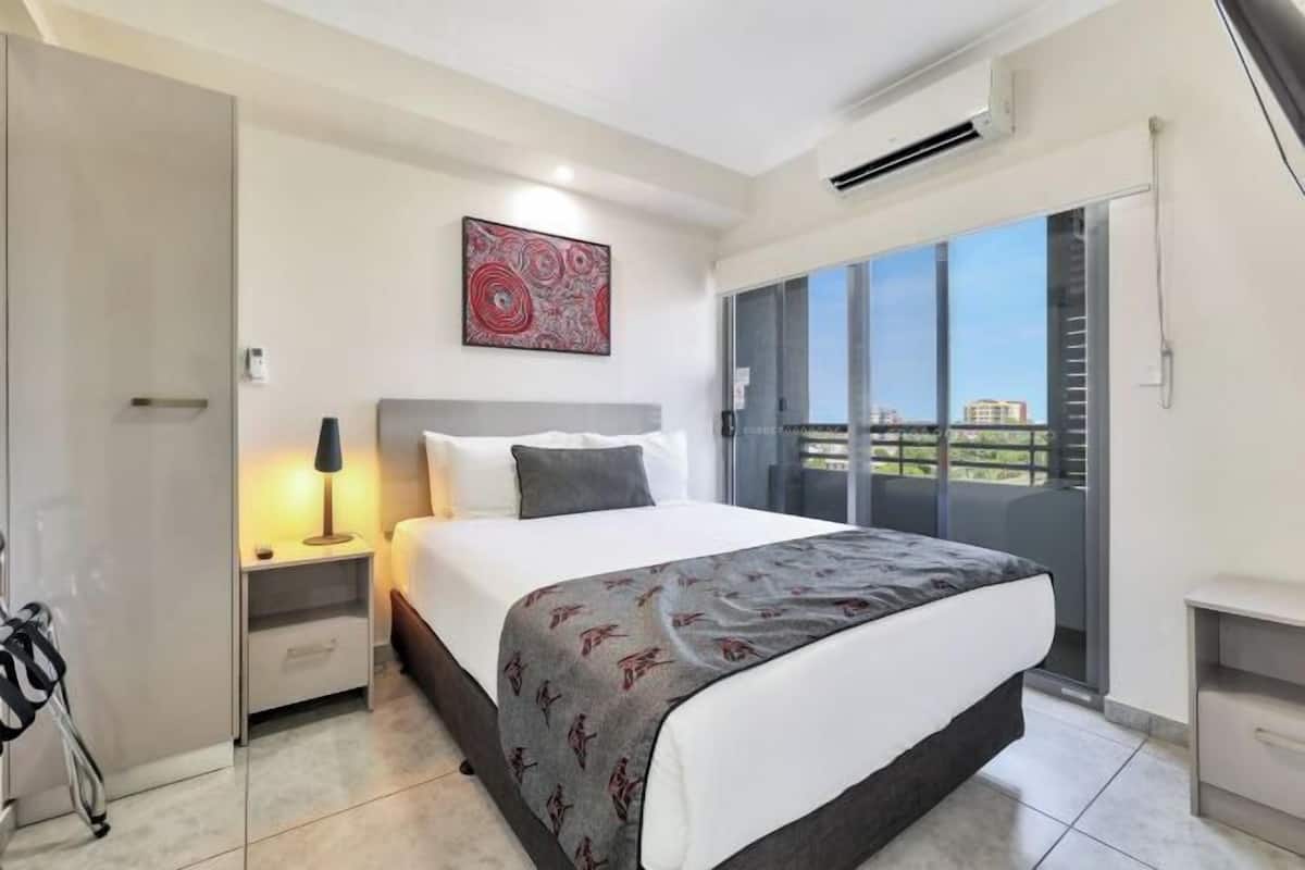 A modern hotel room with a large bed, a nightstand, a lamp, a painting, and a view of the city through a balcony.