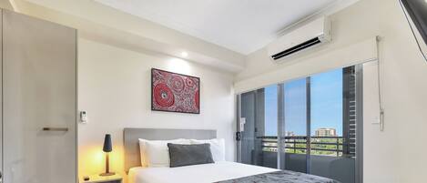 A modern hotel room with a large bed, a nightstand, a lamp, and a view of the city through a balcony.