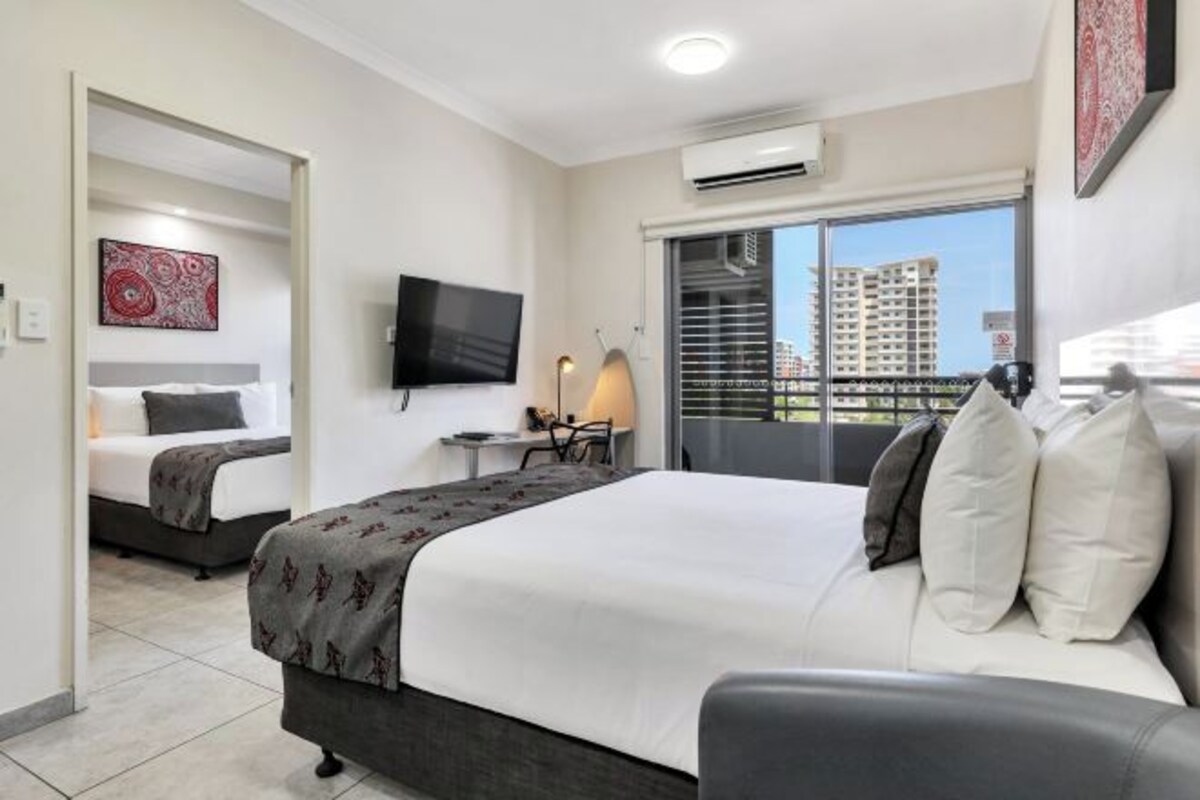 Habitación de hotel con cama, escritorio, silla, televisor y balcón con vistas a edificios.